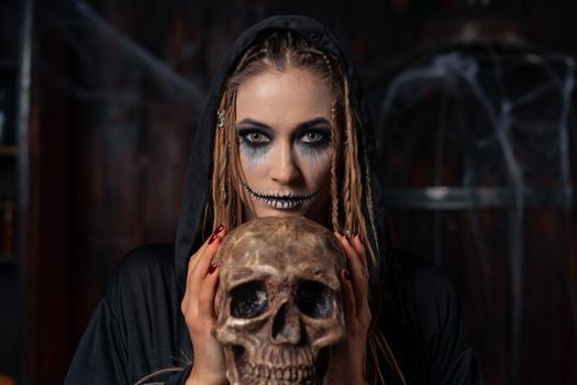 Halloween concept. Witch portrait close up with dreadlocks looking camera dressed black hood standing dark room with cage on background