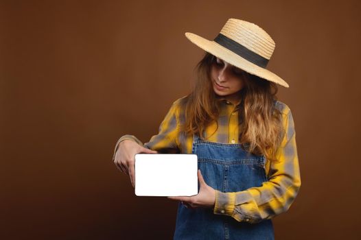Studio portrait. Long-haired Caucasian attractive young woman in country clothes, denim overalls and shirt holds an electronic tablet with a cut-out screen.