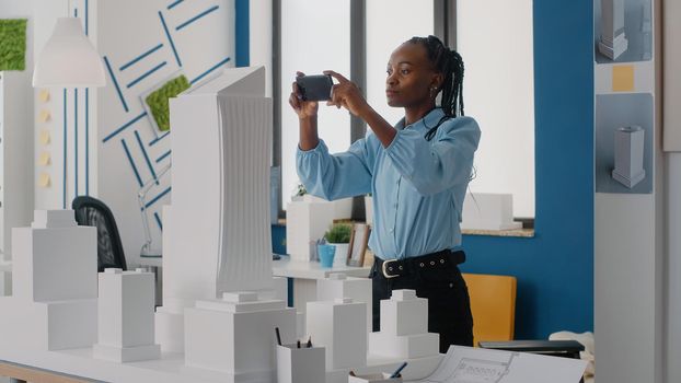 Woman engineer using smartphone to plan layout of building model and maquette