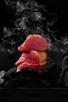 Slices of grilled filet mignon on charcoals on black background in white smoke