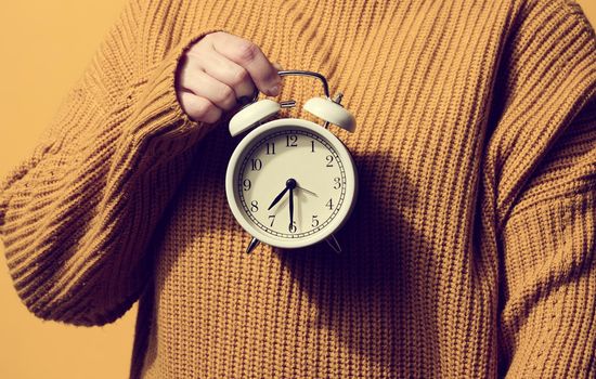round alarm clock in a woman's hand, the time is half past seven in the morning. Woman in orange sweater