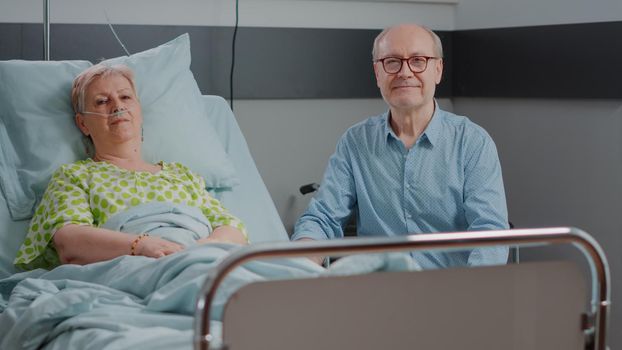 Portrait of senior people sitting in hospital ward, elder woman with disease laying in bed and husband giving support and help. Old person assisting pensioner with sickness at clinic.