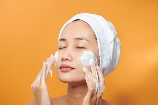 Gentle half-naked woman wrapped in towel washing her face with foaming cleanser isolated over orange background