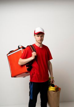 Delivery man in red uniform with a thermal bag on a shoulder holding paper bag with groceries. Food delivery service.