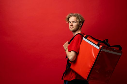 Young courier with thermal bag on red background, space for text. Food delivery service. Delivery guy in a red t-shirt uniform work as courier and holds red thermal food backpack. Service concept.