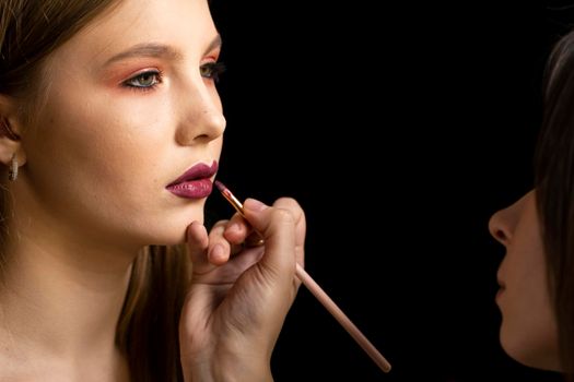 Makeup artist applies red lipstick on a beautiful woman face. Hand of make-up master, painting lips of young beauty model girl. Make up in process.