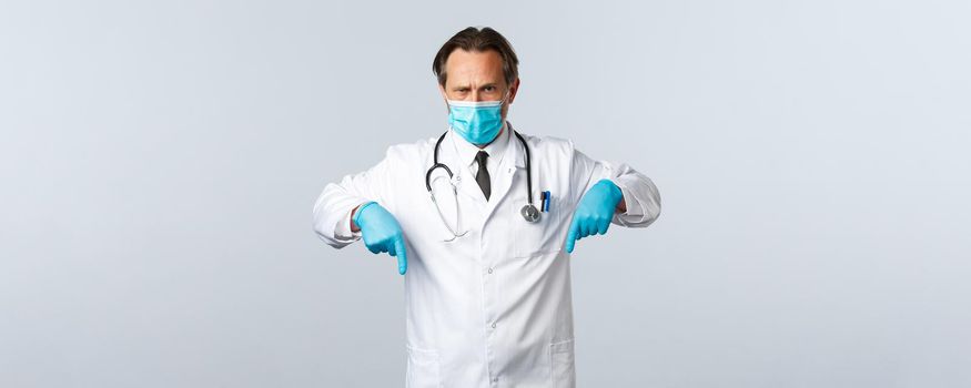 Covid-19, preventing virus, healthcare workers and vaccination concept. Serious determined doctor showing measures against coronavirus, wear medical mask and gloves, pointing fingers down