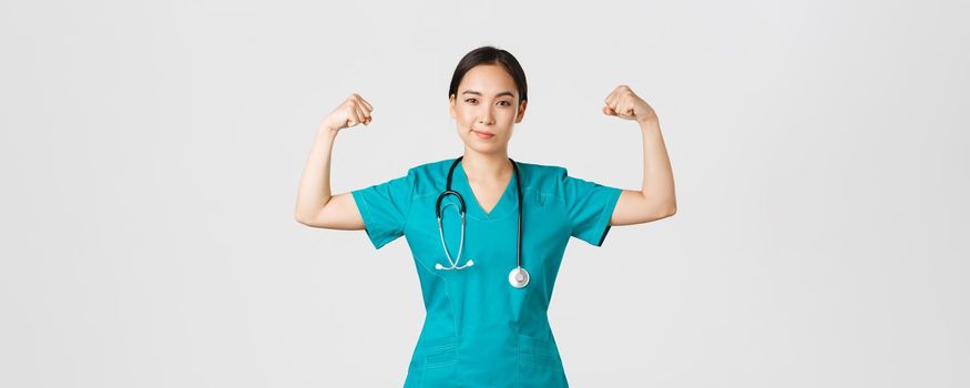 Covid-19, healthcare workers and preventing virus concept. Confident professional asian female doctor, nurse in scrubs smiling sassy and flex biceps, being strong, show-off muscles