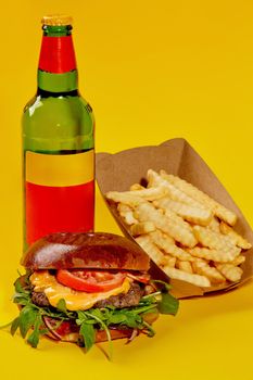Cheeseburger with beef patty, tomatoes, onion and greens with French fries and bottle drink isolated on yellow background