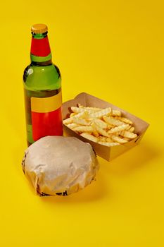 Hamburger in paper wrapping, fries in carton box and bottle of drink on yellow background