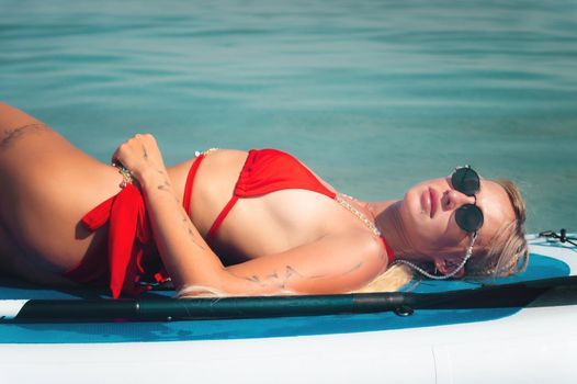 Young sexy surfer woman in red swimsuit and sunglasses lying on her sup board. Relax on stand up paddle on a sunny day Camping concept