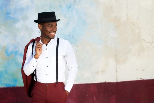 happy young black man with suspenders and hat smiling by wall