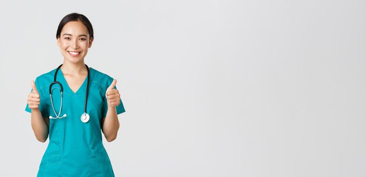 Covid-19, healthcare workers, pandemic concept. Professional confident smiling doctor, female physician in scrubs showing thumbs-up with assured expression, ensure all good, perfect or excellent