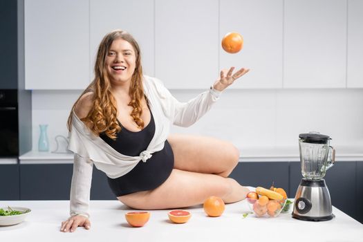 Girl with obese problem. Young sexy chubby white girl in black swimsuit, white shirt at modern kitchen table. juggling fruits in hand. Trying to loose weight fast. Fat barefoot girl low carb diet
