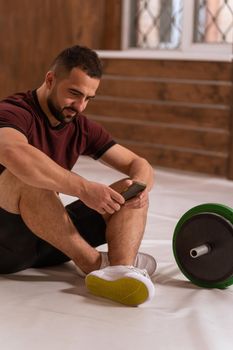 Handsome man sitting on a floor with smartphone in his hands and black and green tone fitness barbell, equipment for weight training concept. Sports equipment for training. Healthy lifestyle concept