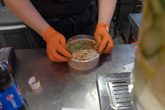 Worker wearing gloves packs tom yum soup into container. Asian restaurant. June 2020. Kyiv, Ukraine