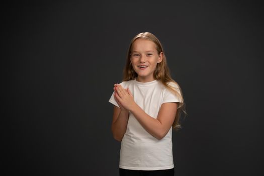 Excited little girl inspired by parents with hands put together happily looking at the camera wearing white t-shirt and black pants isolated on black background