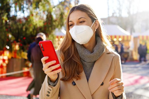 Portrait of elegant woman wearing FFP2 KN95 protective mask using smartphone with people walking in the Christmas markets on the background
