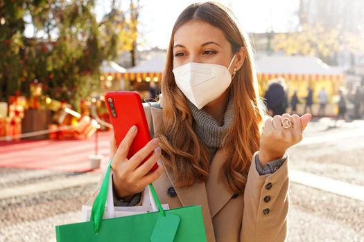 Excited young woman wearing medical mask FFP2 KN95 watching smartphone and holding shopping bags walking in the Christmas Markets