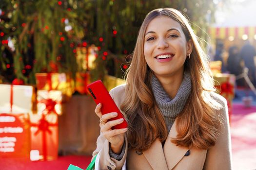 Portrait of an excited pretty young woman holding smartphone and looking at camera with Christmas tree and gifts on background outdoors