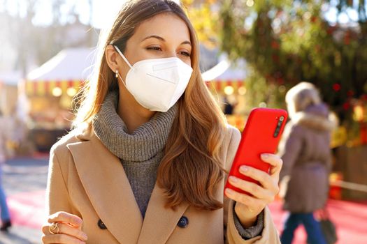 Beautiful young woman wearing medical face mask using smartphone on Christmas time outdoors