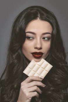 Sensual beautiful brunette young woman bite white bubbly chocolate wearing black jacket with beautiful long dark hair isolated on grey background. Young female model with professional make-up