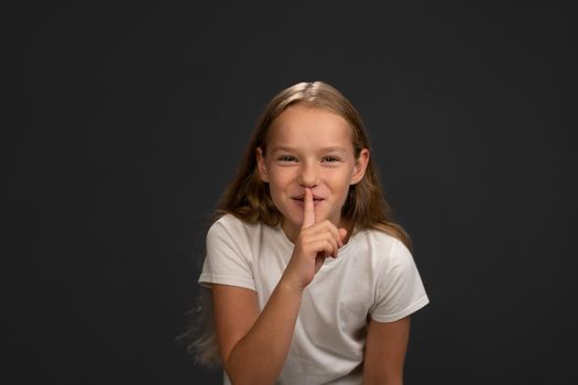 Saying to keep silence little girl holds her finger on her lips telling everyone. Child wearing white t shirt smiling at the camera isolated on dark grey or black background