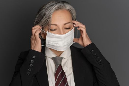 Grey-haired middle-aged business woman putting on a protective mask with eyes closed isolated on grey background. Portrait of modern senior woman in studio wearing business clothes