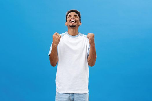 African american man being happy finally gaining success reaching victory raising clenching teeth rejoicing from happiness smiling joyfully raising head up celebrating triumph over blue background