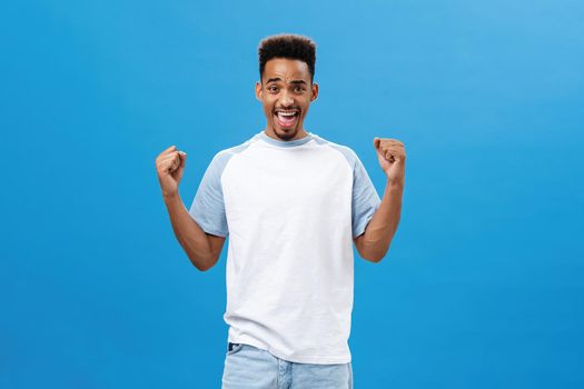 Supportive and cheering african american young sportsman in trendy t-shirt raising fists in victory and triumph gesture yelling yes with delight and satisfaction celebrating, receiving positive result