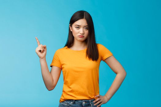 Moody upset asian girlfriend, sulking look offended and displeased pointing upper left corner, pouting disappointed, standing distressed blue background wear yellow t-shirt