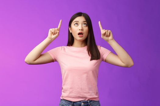 Surprised impressed asian girl brunette look fascinated up, drop jaw gasping astonished, check out impressive interesting promo, pointing top advertisement amazed, stand purple background