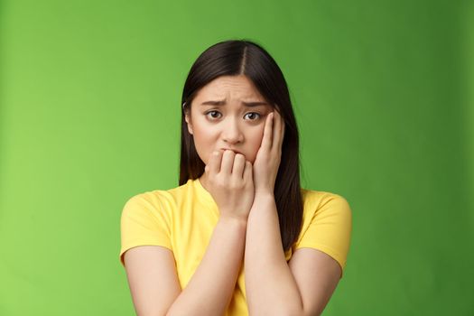 Innocent insecure timid asian scared girl panicking, standing afraid victim terrified, touch cheek shocked, frowning stunned, biting fingernails, anxiously stare camera, green background