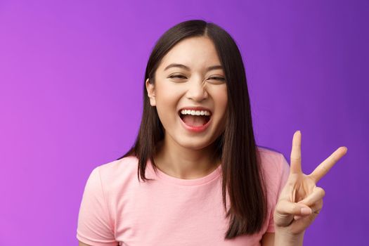 Headshot sincere carefree laughing asian girl having fun, show peace victory sign, joyfully look camera, express enthusiasm and happiness, enjoy funny friendly company, stand purple background