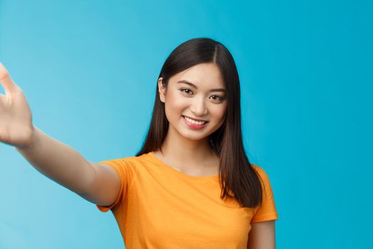 Close-up attractive friendly modern vietnamese girl extend hand, taking selfie, smiling joyfully, make photograph smartphone, hold camera arm, stand blue background joyful video-call friend