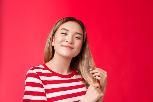 Close-up satisfied cute carefree asian blond girl feeling delighted and pleased after haircare procedure touch healthy hair tilt head smiling broadly like new hairdresser, stand red background