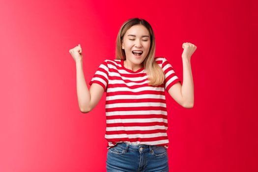 Hooray I did it. Triumphing cute cheerful asian blond girl make fist pumps closed eyes happy broad smile, celebrating accomplishment feel satisfaction finally winning achieve success red background