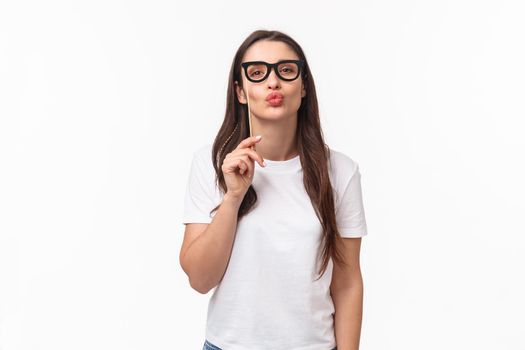 Entertainment, fun and holidays concept. Portrait of silly and cute young attractive girl wearing carnaval mask glasses, folding lips to give air kiss, pouting and standing white background