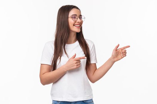 Waist-up portrait of cheerful young female student, programmer, pointing fingers and looking at right copy space, smiling and laughing as seeing something entertaining and funny, white background