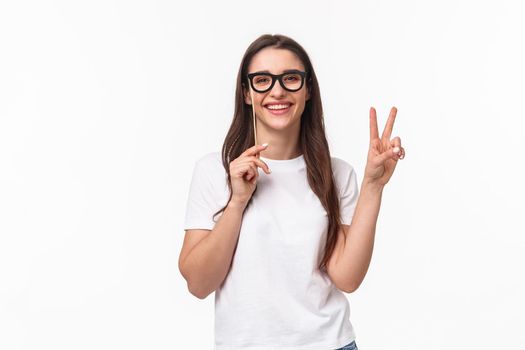 Entertainment, fun and holidays concept. Portrait of carefree cheerful young girl enjoying awesome party, celebrating show peace sign and wearing carnaval mask, standing white background