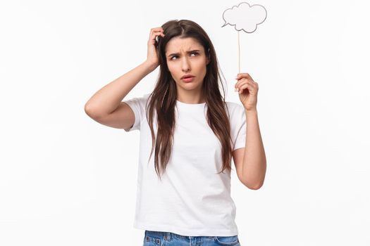 Entertainment, fun and holidays concept. Portrait of puzzled, indecisive young brunette girl, female student calculating something in mind, hold comment cloud stick near head, scratching and thinking