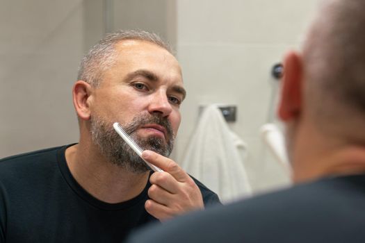 Middle-aged handsome man brushing his beard in the morning in bathroom looking in mirror