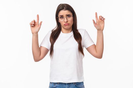 Waist-up portrait of distressed, sad and gloomy indecisive young brunette woman in t-shirt and glasses, shrugging pointing fingers up and making miserable uneasy face, dont know, cant decide