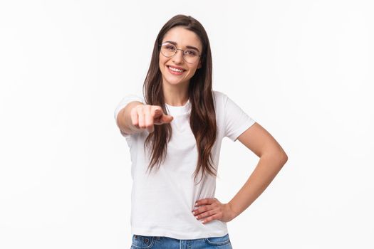 Waist-up portrait of happy smiling young woman inviting you join her team, recruit person, pointing finger at camera grinning and standing confident in her choice, white background