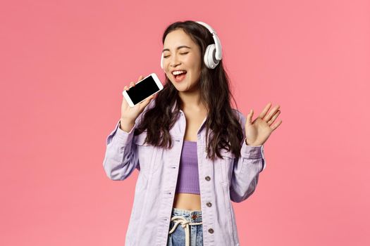 Waist-up portrait of stylish happy woman using headphones to listen awesome new track, download karaoke app and using smartphone like microphone, singing along awesome music