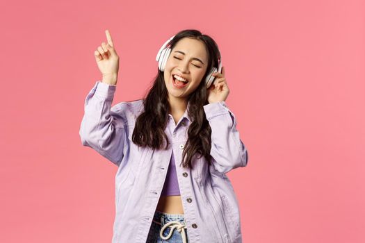 Portrait of attractive carefree asian girl chilling and vibing, listening music in headphones, raising finger up while trying sing along and reach highest note in song, pink background