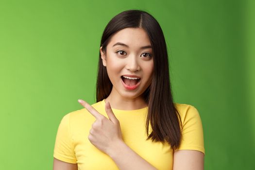 Enthusiastic impressed cute asian brunette describing new haircare product features amused and amazed, lively talking poining left astonished, have interesting discussion, stand green background