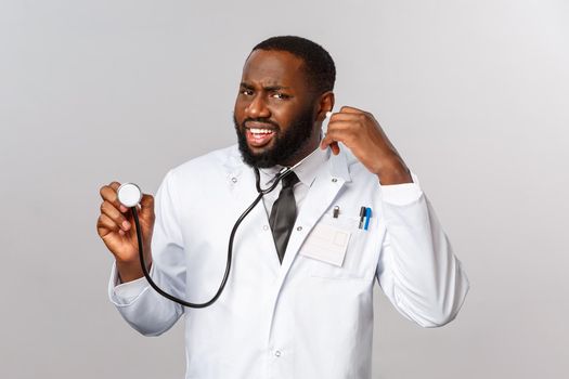 Covid19, hospital check-up and healthcare concept. Portrait of confused grimacing african-american doctor cant hear anything as patient talking during checking his lungs with stethoscope