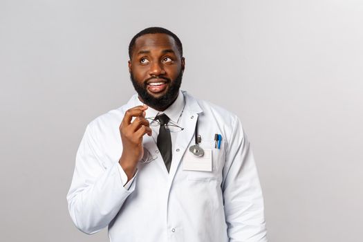 Healthcare, medicine and hospital treatment concept. Thoughtful handsome young doctor, african-american therapist thinking what order for food at lunch, bite glasses rim and look up daydreaming