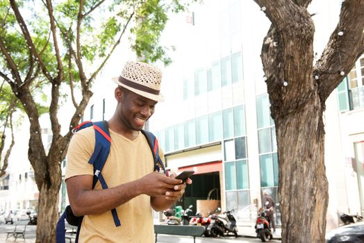 happy african american man with bag and hat walking with mobile phone outdoors